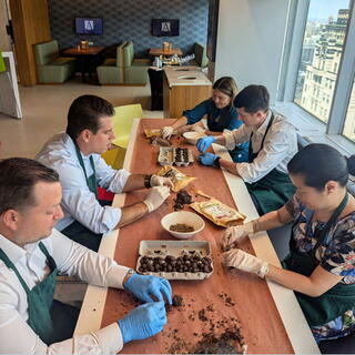 Five people wearing green aprons and latex gloves are seated around a long table in a high-rise office breakroom, engaged in a hands-on team-building activity. They are using soil and organic materials to create small round seed balls, which are being placed into cardboard egg cartons. The room features modern furniture, booth seating, and large windows that offer an elevated view of a city skyline with historic architecture.